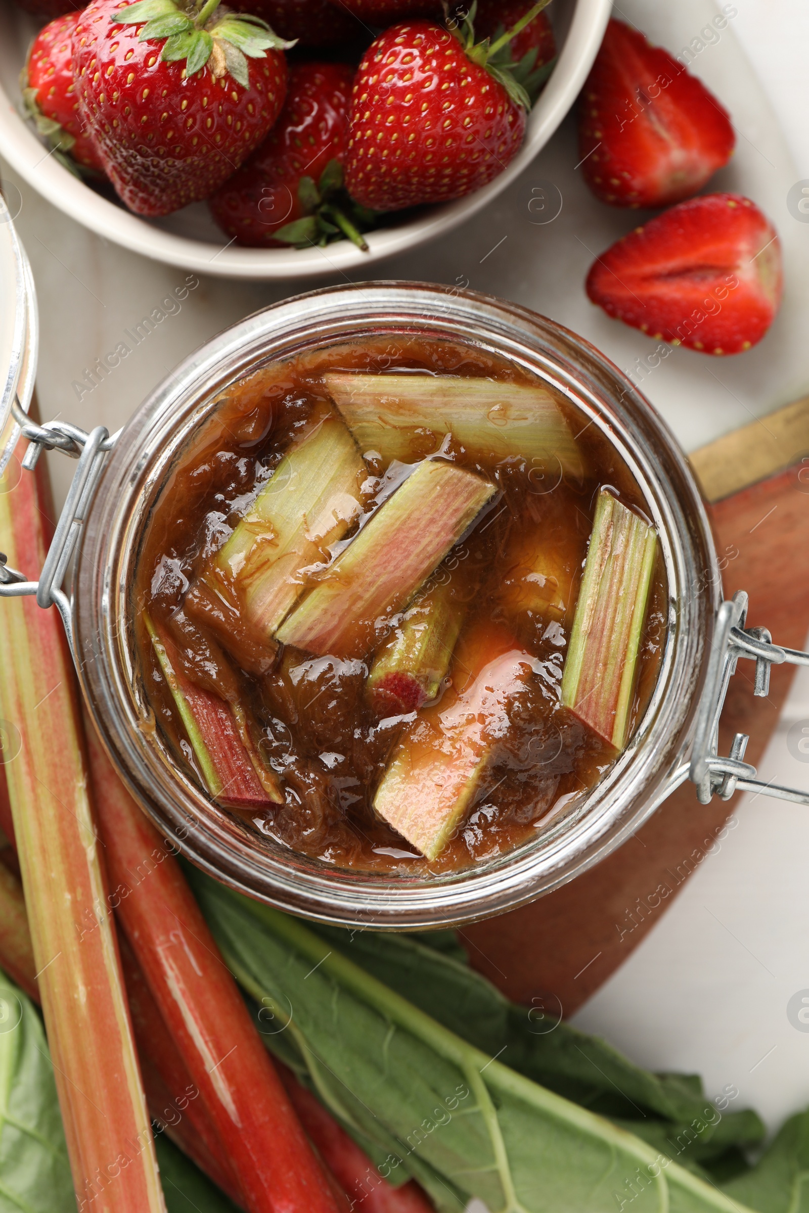 Photo of Jar of tasty rhubarb jam, fresh stems and strawberries on white table, flat lay