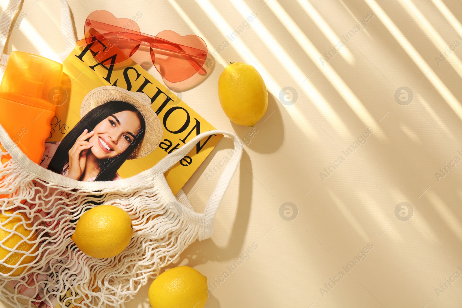 Photo of String bag with fresh lemons, fashion magazine and beach accessories on beige background, flat lay. Space for text