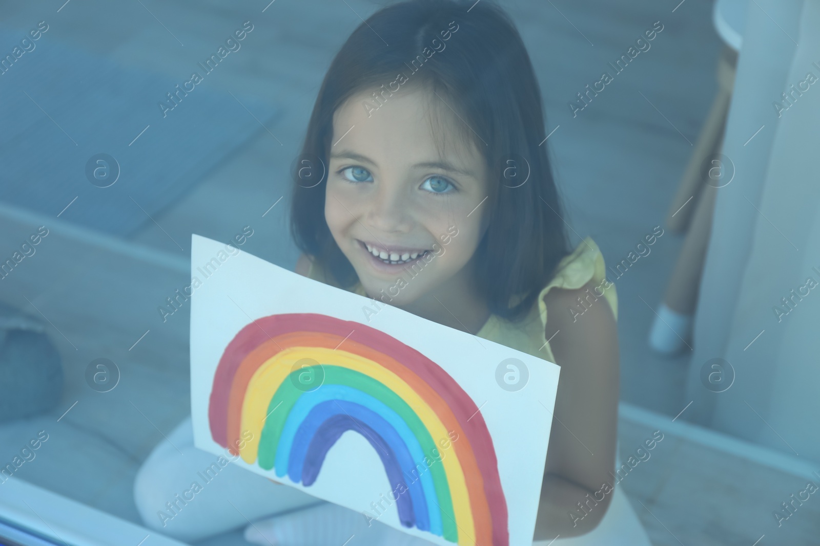 Photo of Little girl with picture of rainbow near window, view from outdoors. Stay at home concept