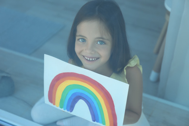 Little girl with picture of rainbow near window, view from outdoors. Stay at home concept