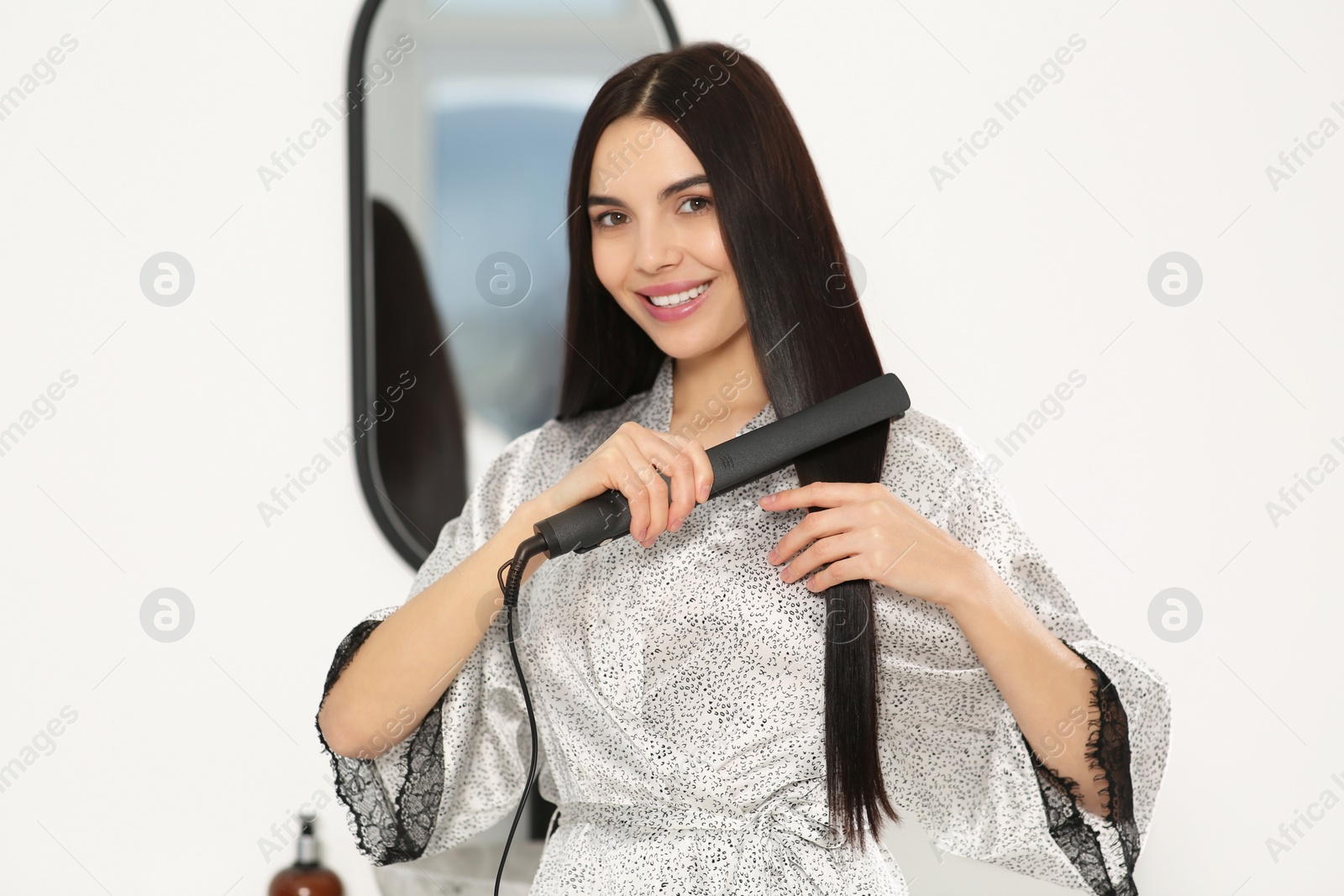 Photo of Beautiful happy woman using hair iron near mirror indoors