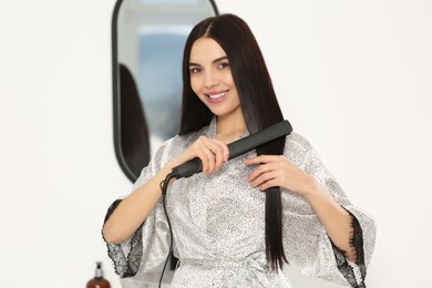 Beautiful happy woman using hair iron near mirror indoors