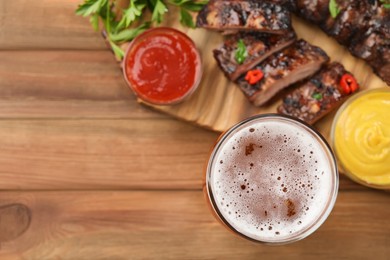 Photo of Glass of beer, tasty grilled ribs and sauces on wooden table, flat lay. Space for text