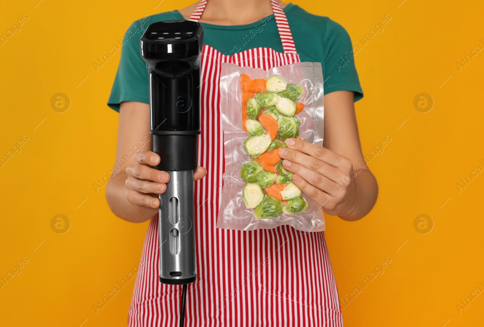 Photo of Woman holding sous vide cooker and vegetables in vacuum pack on orange background, closeup
