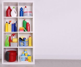 Photo of Shelving unit with detergents and cleaning tools near white wall indoors