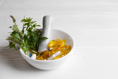 Photo of Mortar with fresh green herbs and pills on white wooden table, space for text