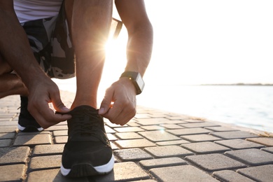 Photo of Man with fitness tracker tying shoelaces near river, closeup. Space for text