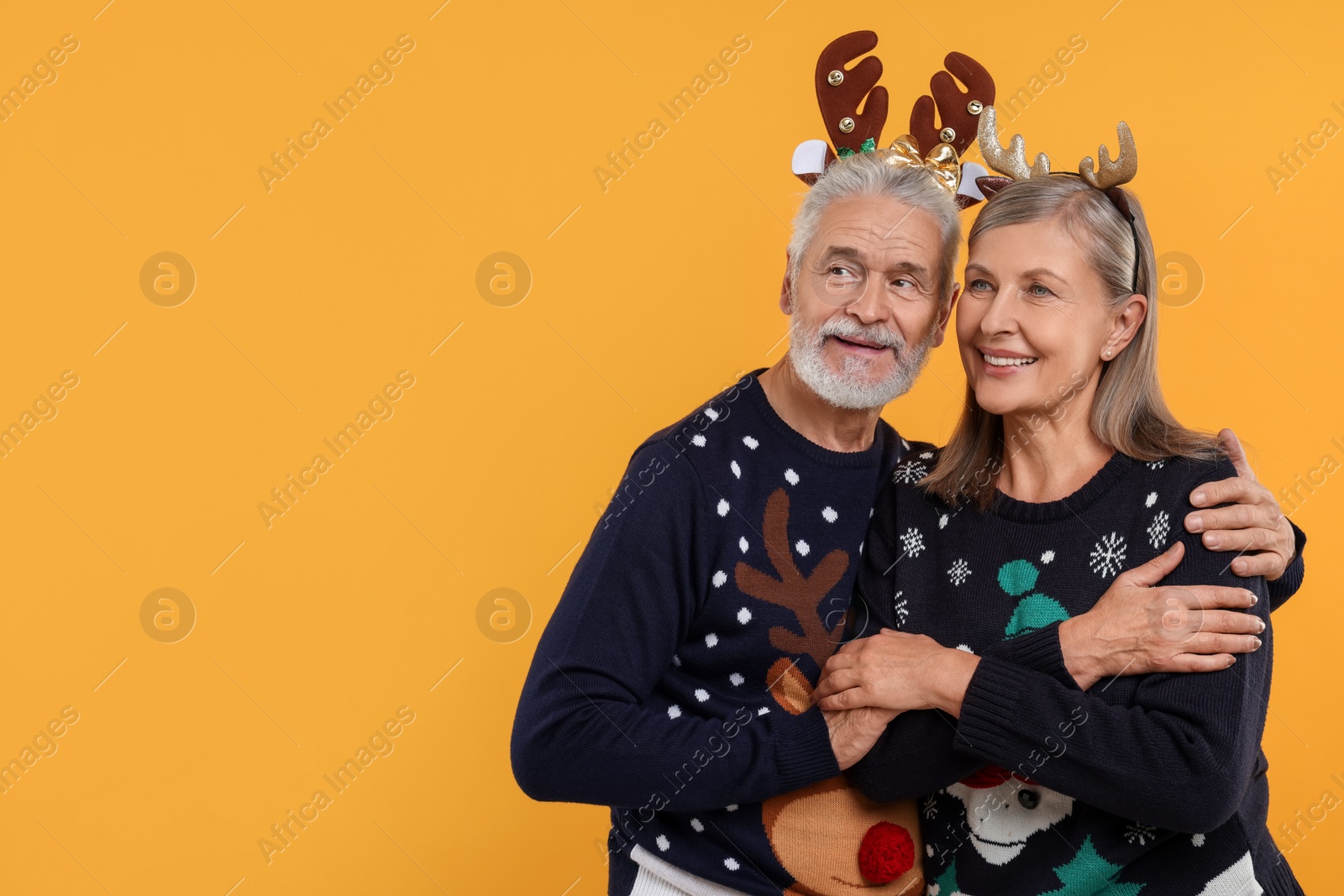 Photo of Senior couple in Christmas sweaters and reindeer headbands on orange background. Space for text