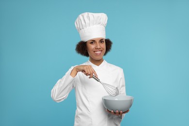 Photo of Happy female chef in uniform holding bowl and whisk on light blue background