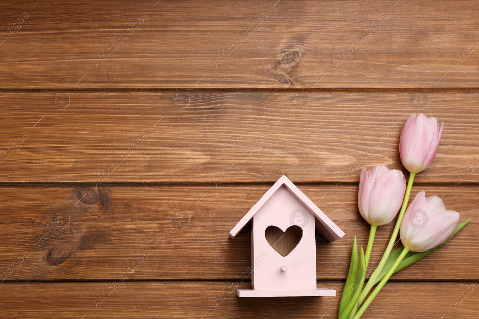 Photo of Beautiful bird house with heart shaped hole and tulips on wooden background, space for text. Spring flat lay composition