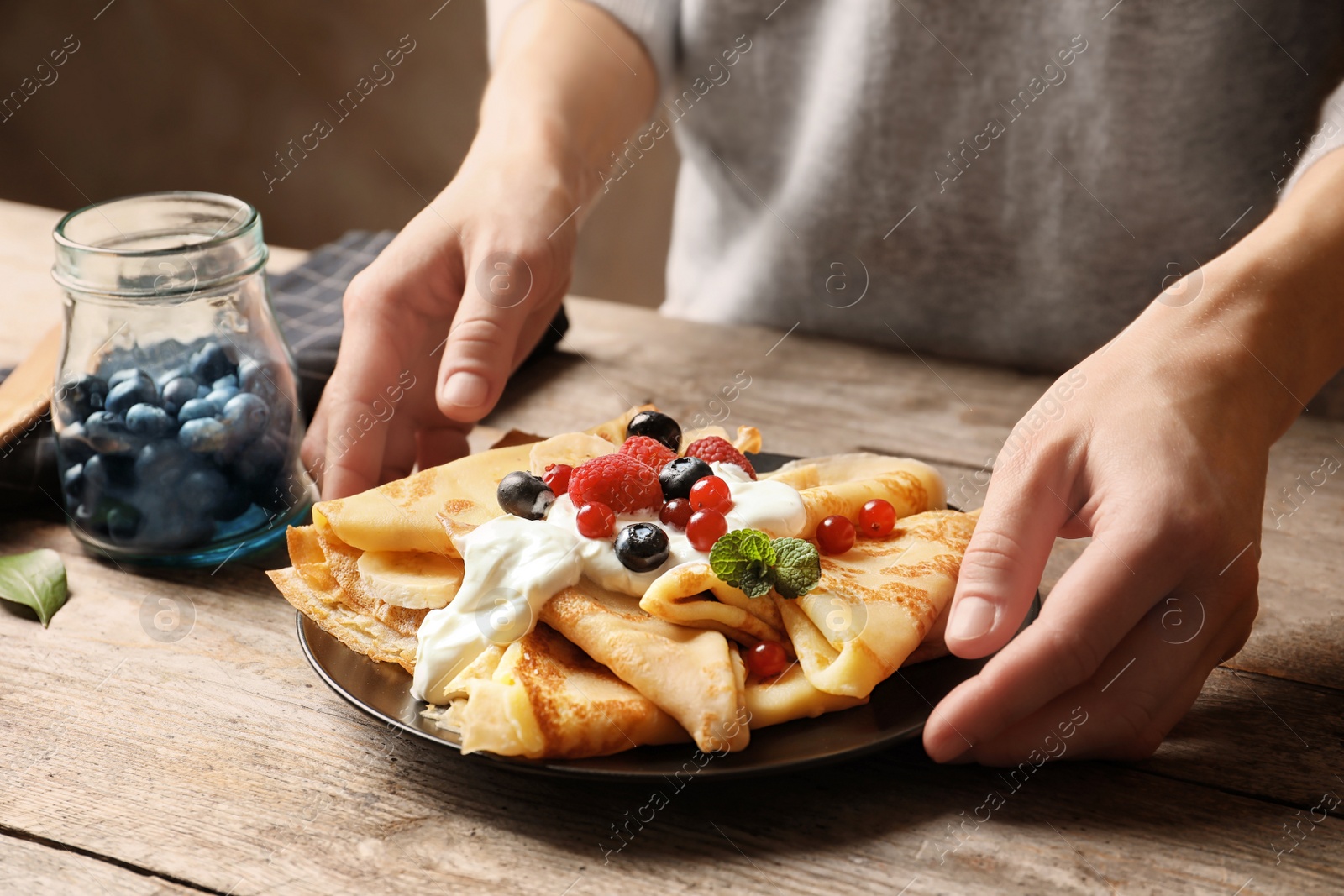 Photo of Woman putting plate with thin pancakes, berries and sour cream on table