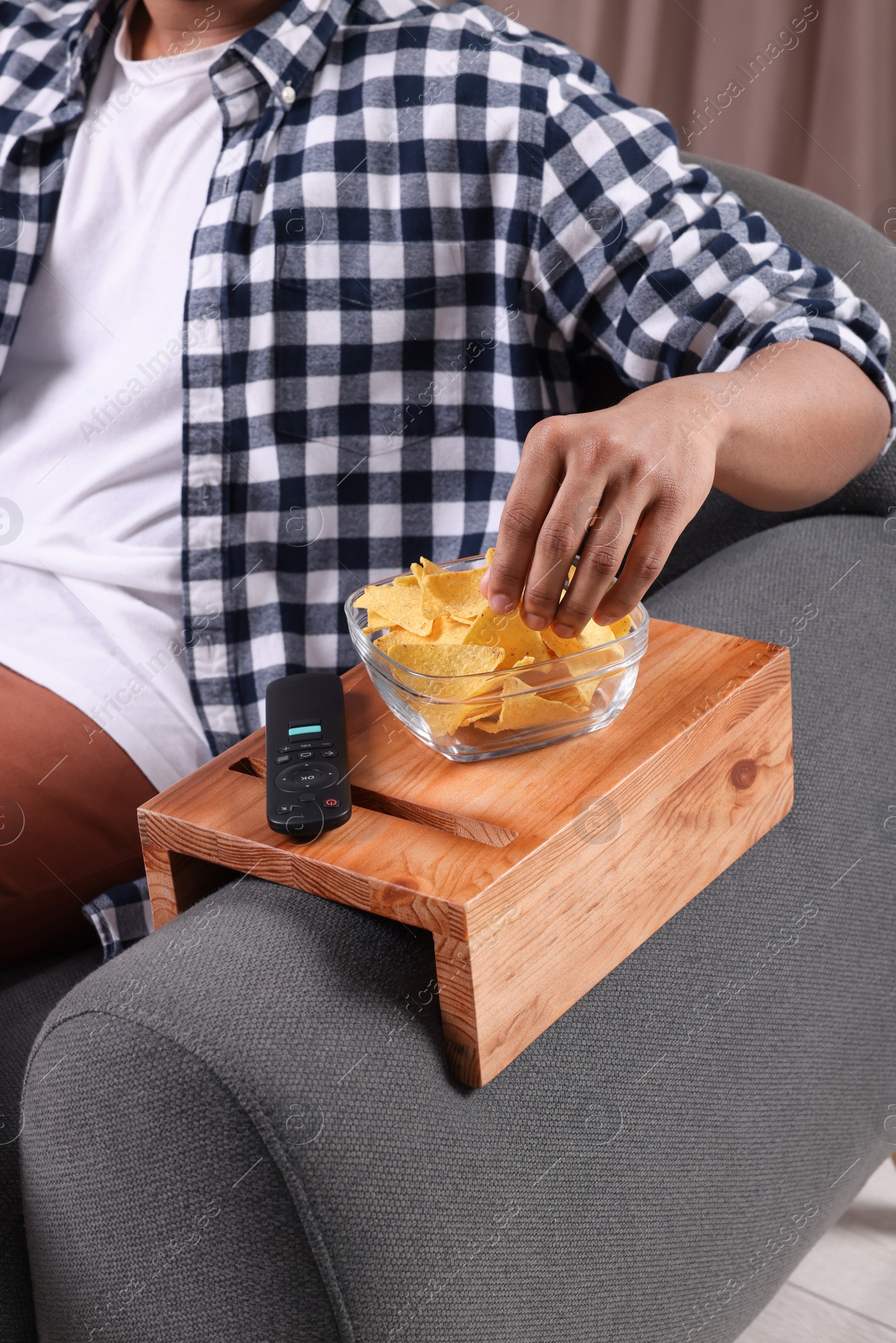 Photo of Man eating nacho chips on sofa with wooden armrest table at home, closeup