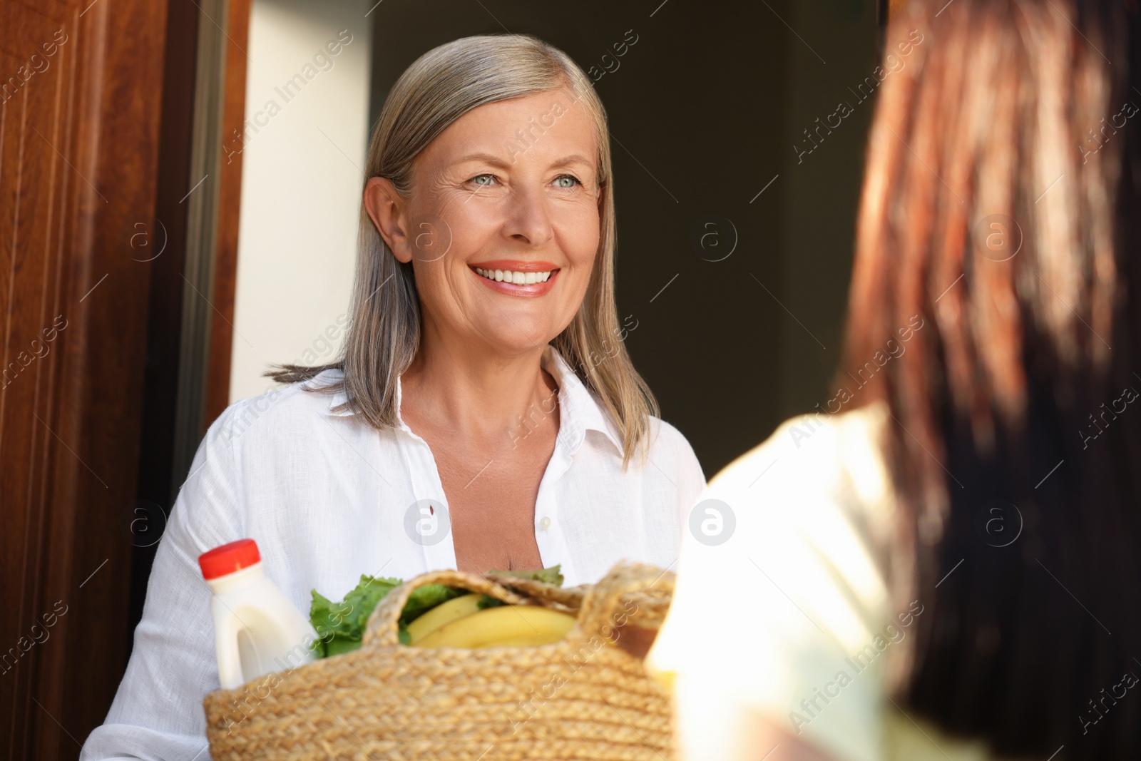 Photo of Young woman with wicker bag of products helping her senior neighbour outdoors