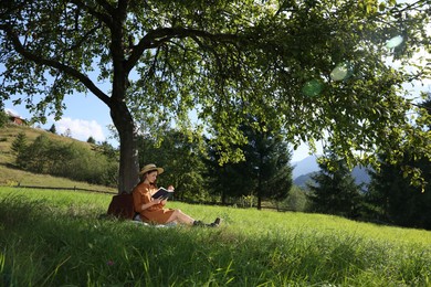 Young woman reading book under tree on meadow in mountains