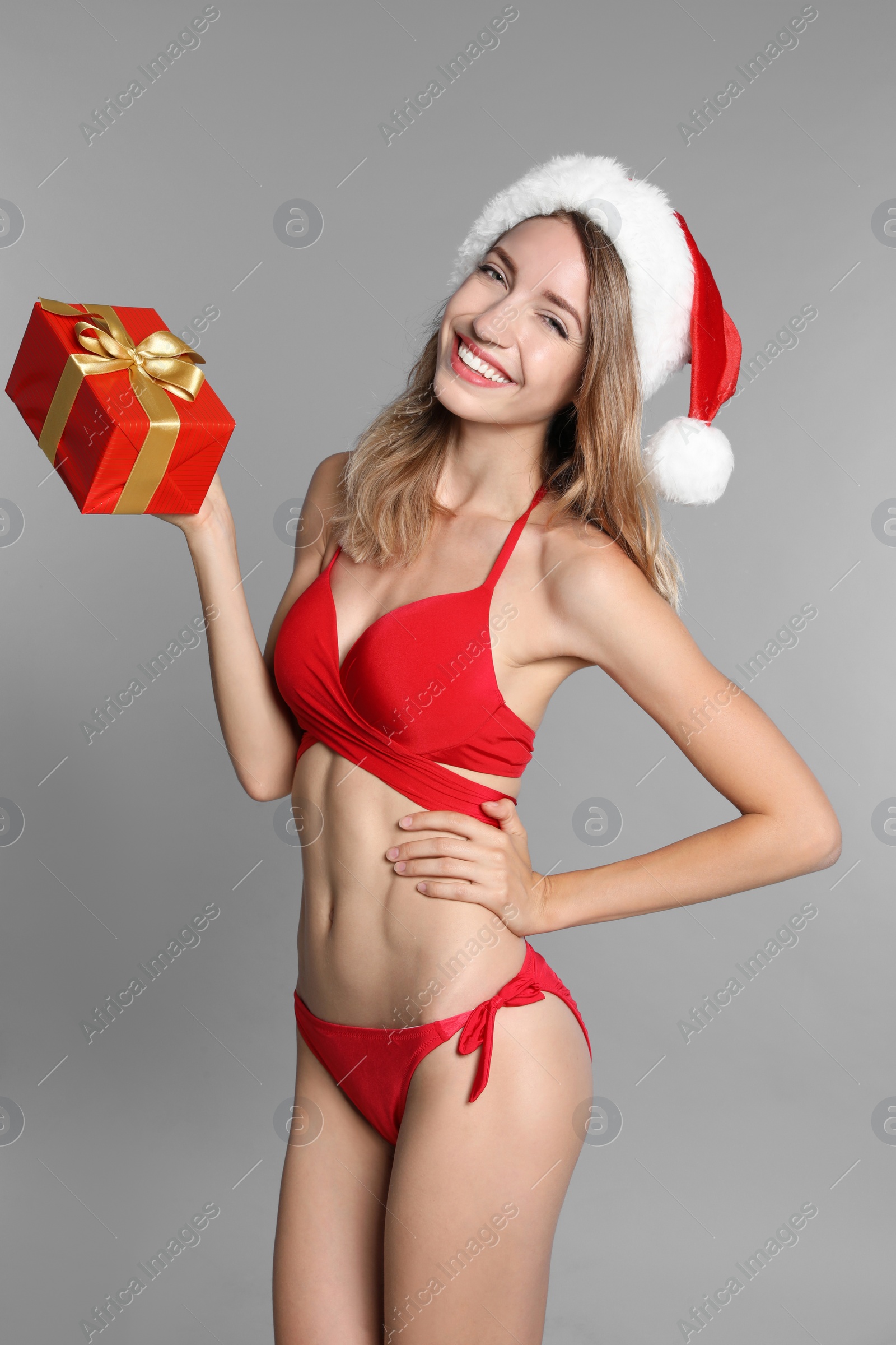 Photo of Happy young woman in red bikini and Santa hat with gift box on light grey background. Christmas celebration