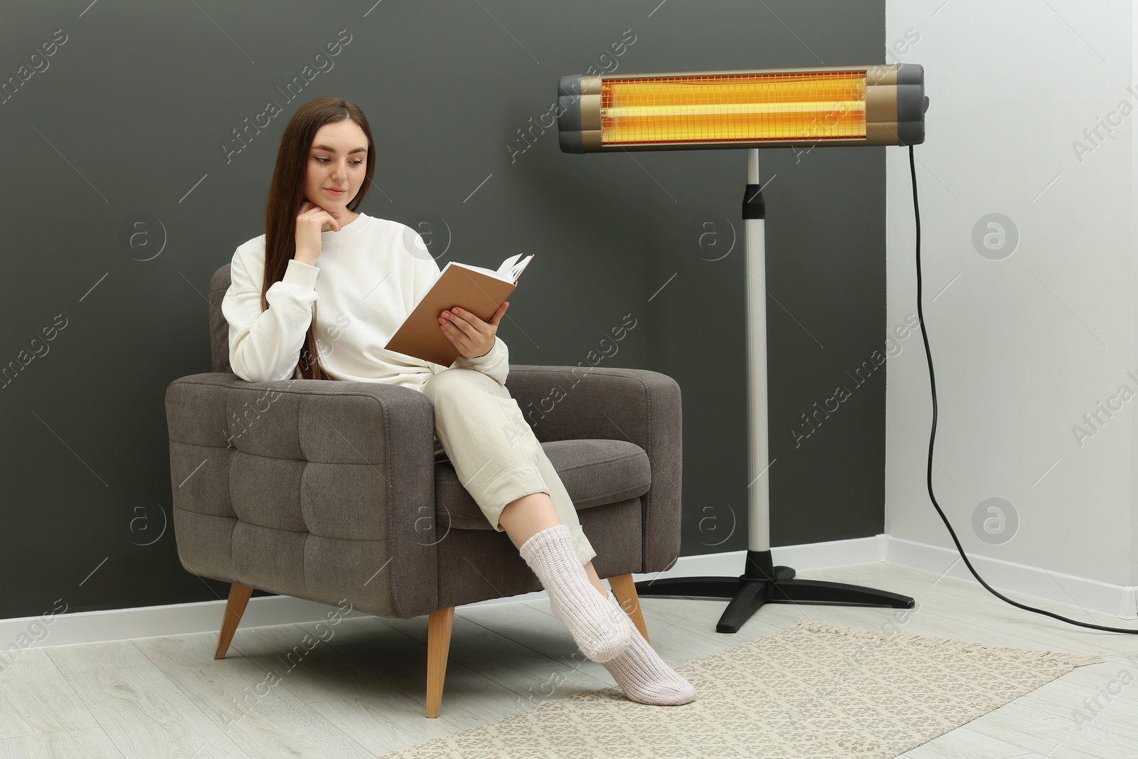 Photo of Young woman reading book on armchair near electric infrared heater at home. Space for text