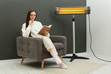 Young woman reading book on armchair near electric infrared heater at home. Space for text