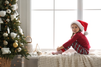Cute little girl in Santa hat on window sill near Christmas tree at home