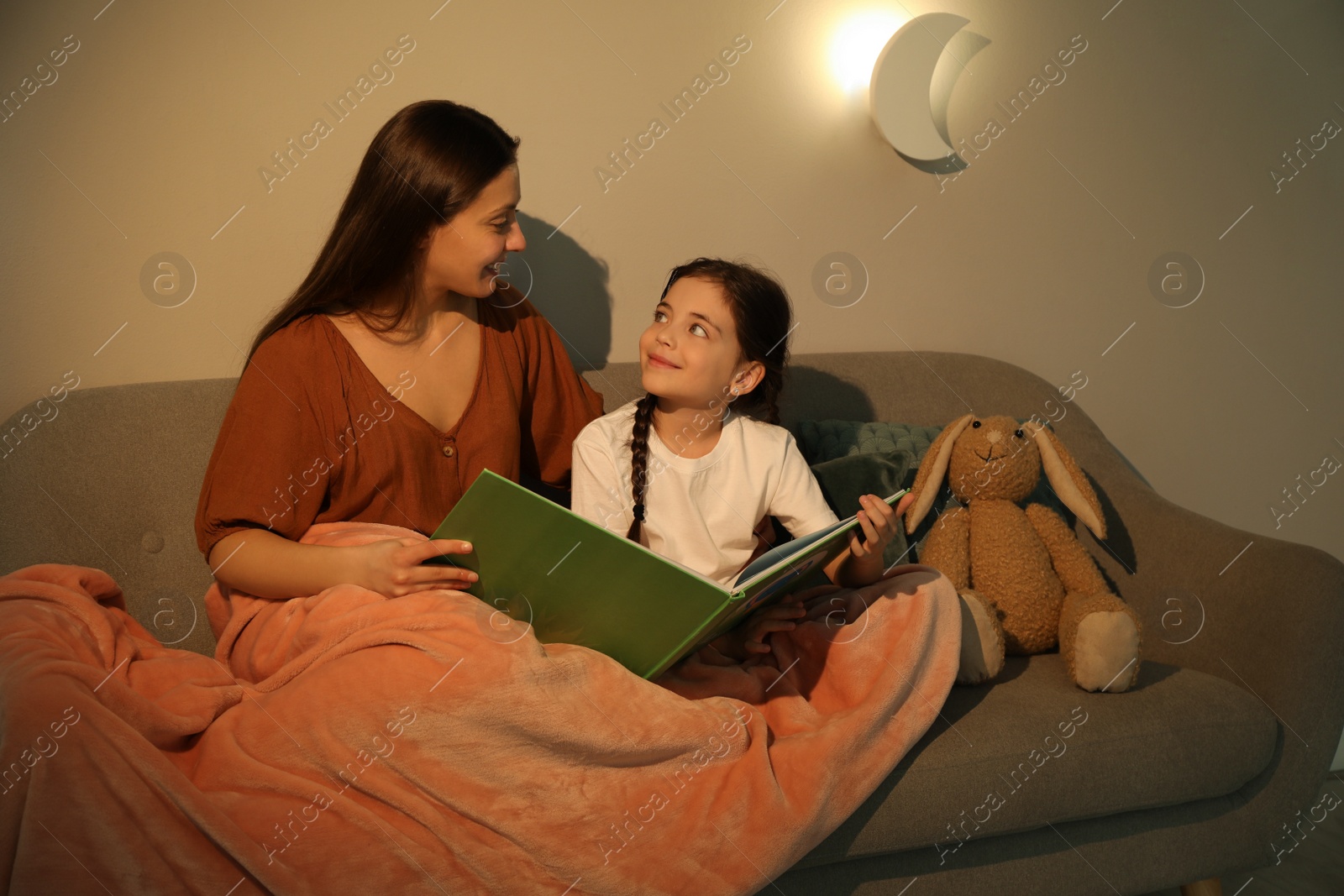 Photo of Little girl with mother reading book in living room lit by night lamp