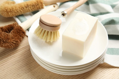 Photo of Cleaning brush, soap bar and plates on wooden table, closeup. Dish washing supplies