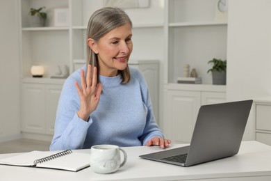 Photo of Happy woman waving hello during video call at table indoors