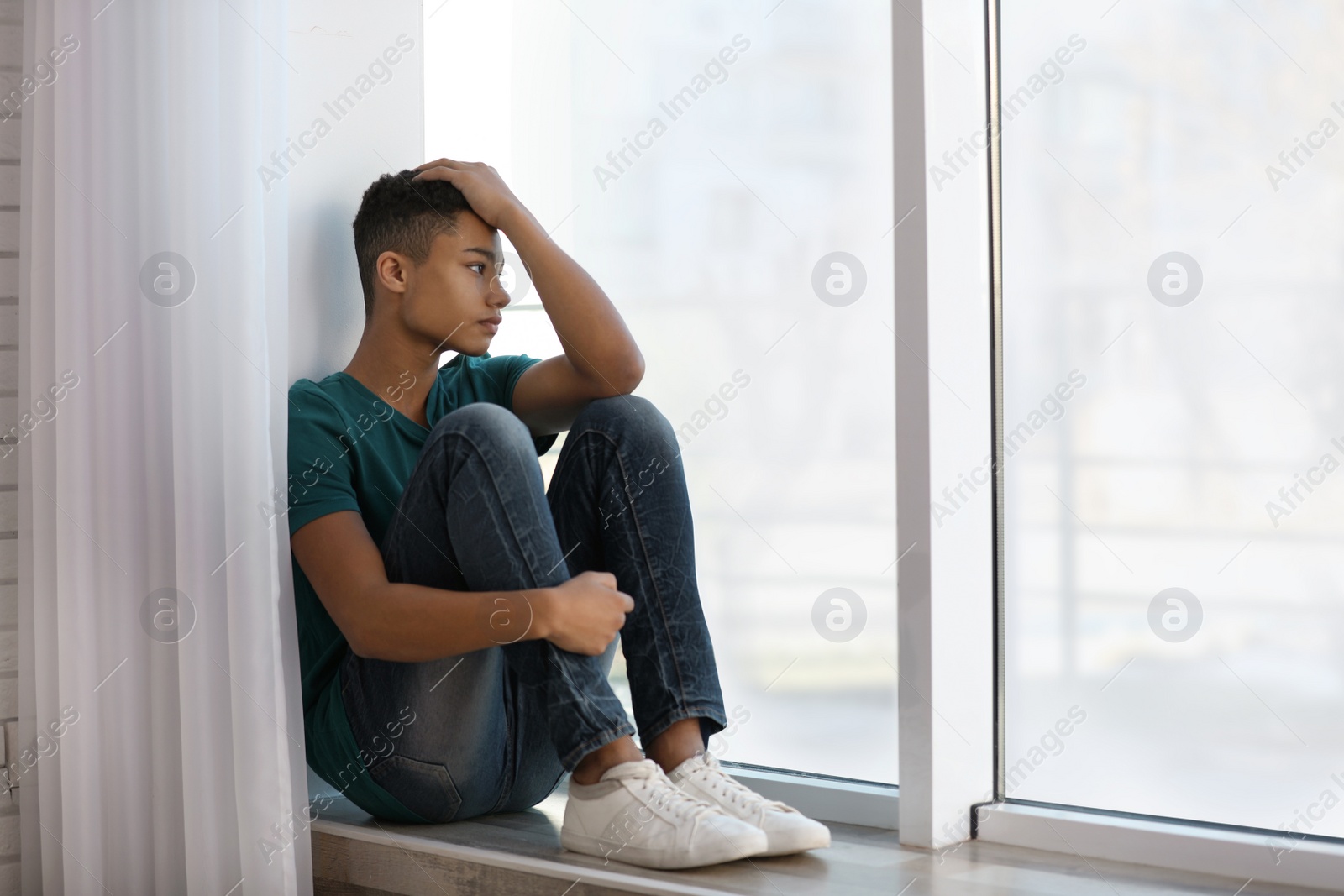 Photo of Upset African-American teenage boy sitting alone near window