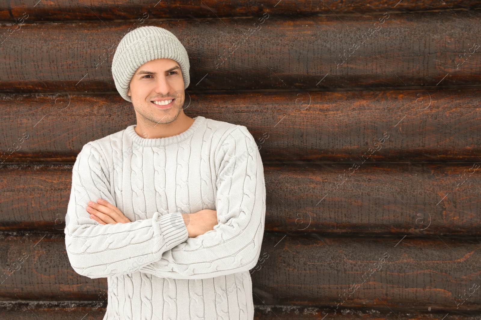 Photo of Handsome man wearing warm sweater and hat near wooden wall, space for text. Winter season