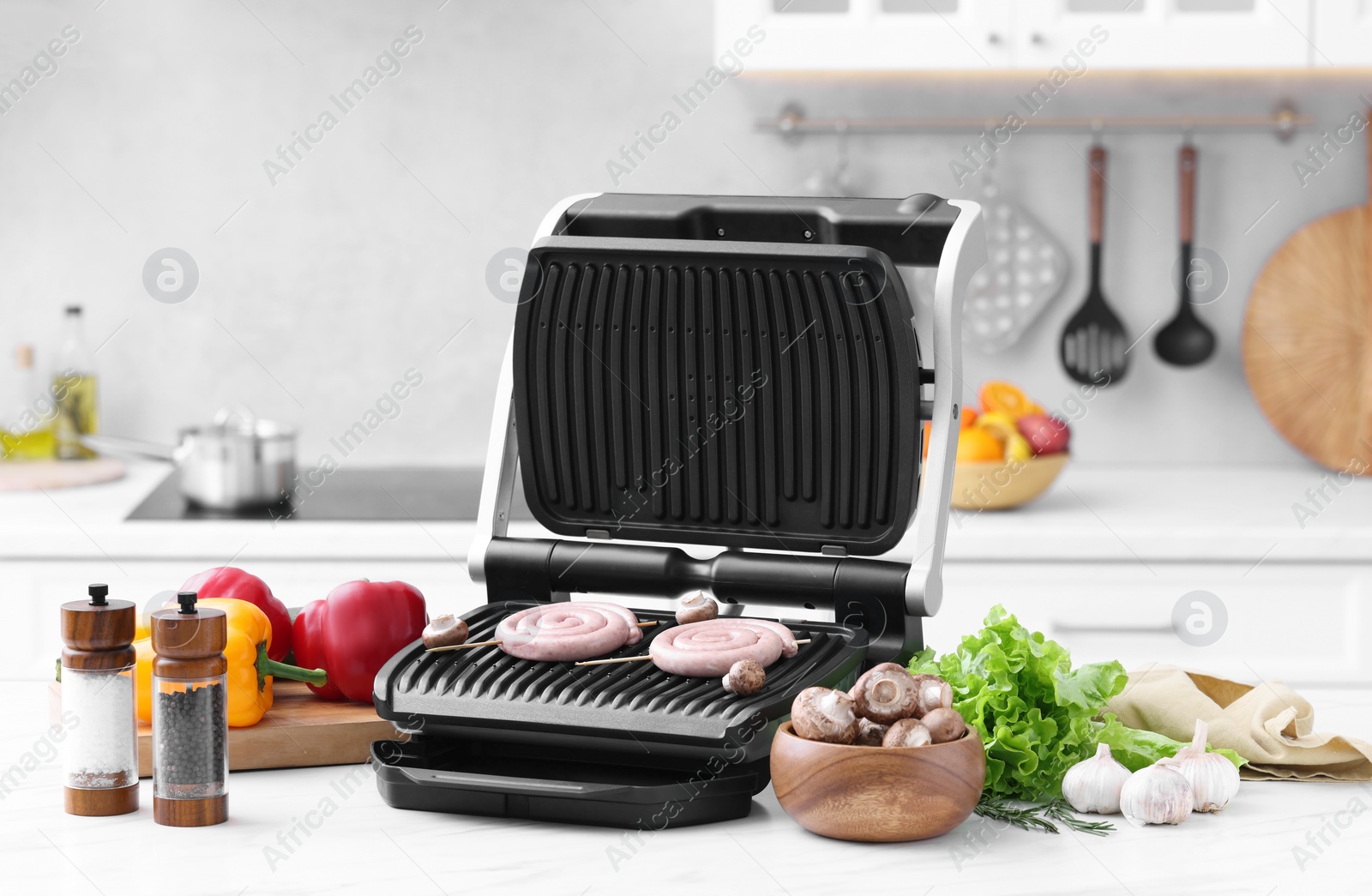 Photo of Electric grill with homemade sausages, mushrooms, vegetables and spices on white marble table in kitchen