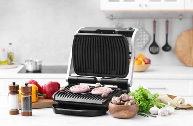 Electric grill with homemade sausages, mushrooms, vegetables and spices on white marble table in kitchen