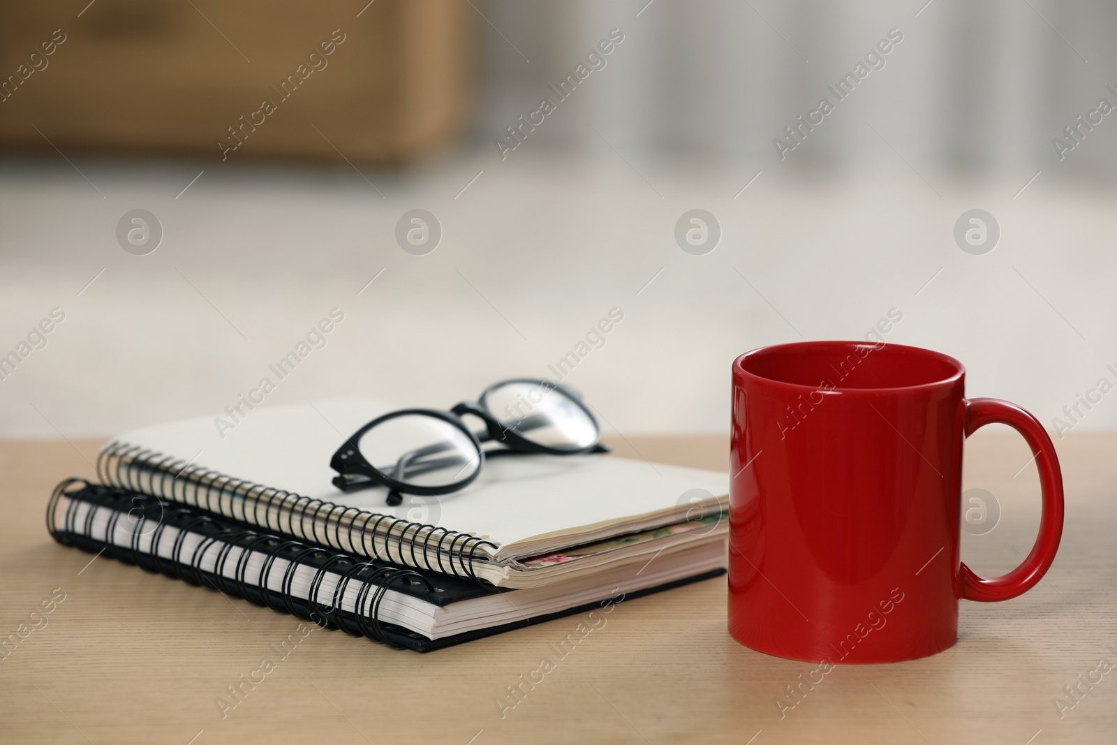 Photo of Red ceramic mug, notebooks and glasses on wooden table at workplace. Space for text