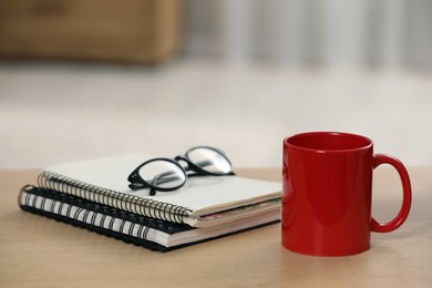 Photo of Red ceramic mug, notebooks and glasses on wooden table at workplace. Space for text