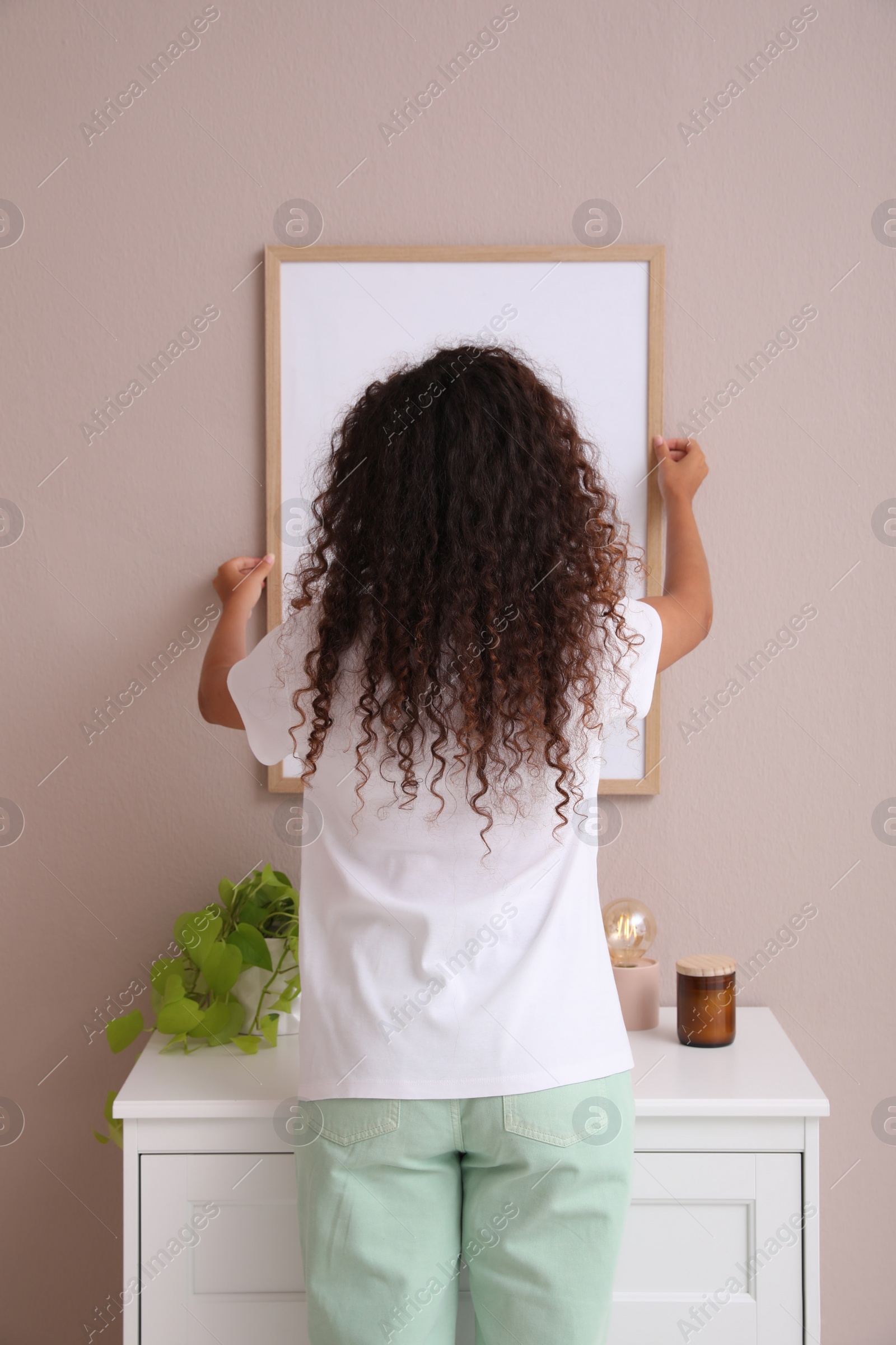 Photo of Woman hanging frame on pale rose wall over chest of drawers in room, back view