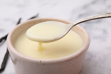 Photo of Condensed milk flowing down from spoon into bowl on white table, closeup