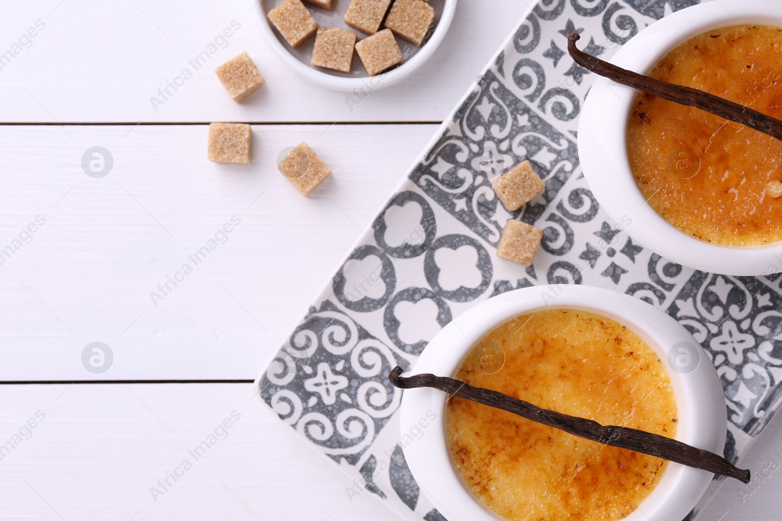 Photo of Delicious creme brulee in bowls, vanilla pods and sugar cubes on white wooden table, flat lay. Space for text