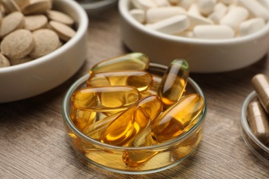 Dietary supplement capsules in bowl on wooden table, closeup