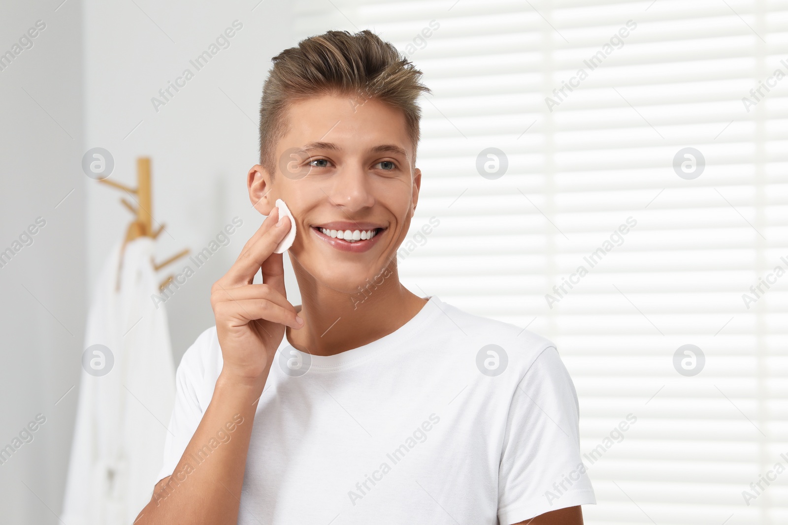 Photo of Handsome young man cleaning face with cotton pad in bathroom
