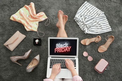 Photo of Woman surrounded by clothes and accessories using laptop with Black Friday announcement while sitting on grey carpet, top view