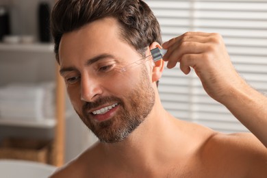 Smiling man applying cosmetic serum onto his face indoors, closeup