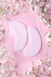 Package with under eye patches and flowers on light pink background, flat lay. Cosmetic product