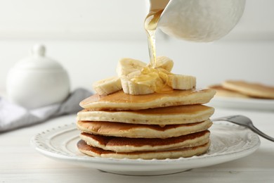 Photo of Pouring honey from jug onto delicious pancakes with bananas at white wooden table, closeup