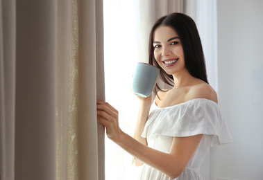 Woman with cup of coffee opening window curtain at home in morning