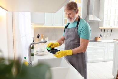 Senior man in protective gloves washing plate above sink in kitchen