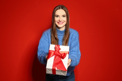 Young woman in Christmas sweater holding gift box on red background