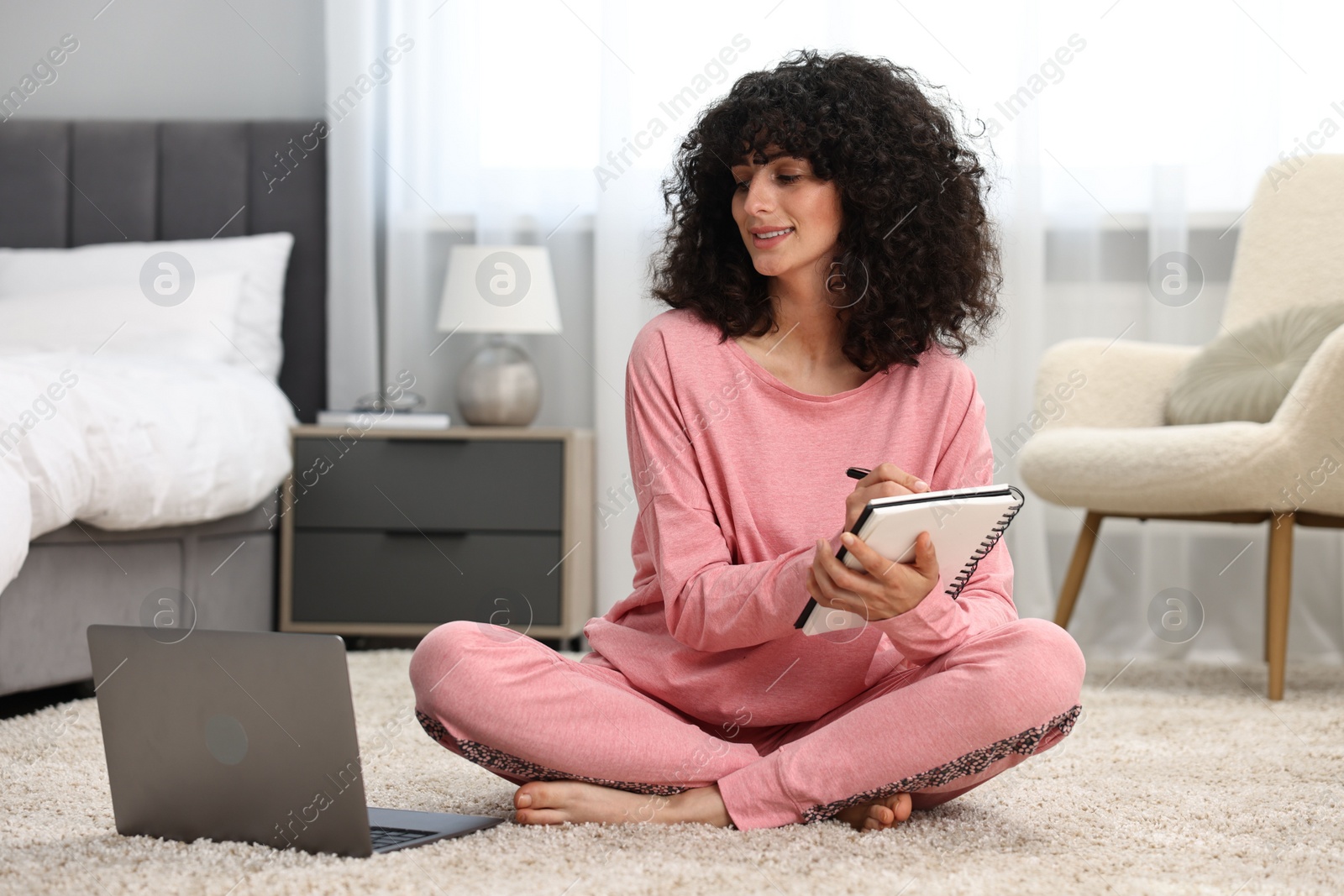 Photo of Beautiful young woman in stylish pyjama with laptop taking notes in notebook on floor at home