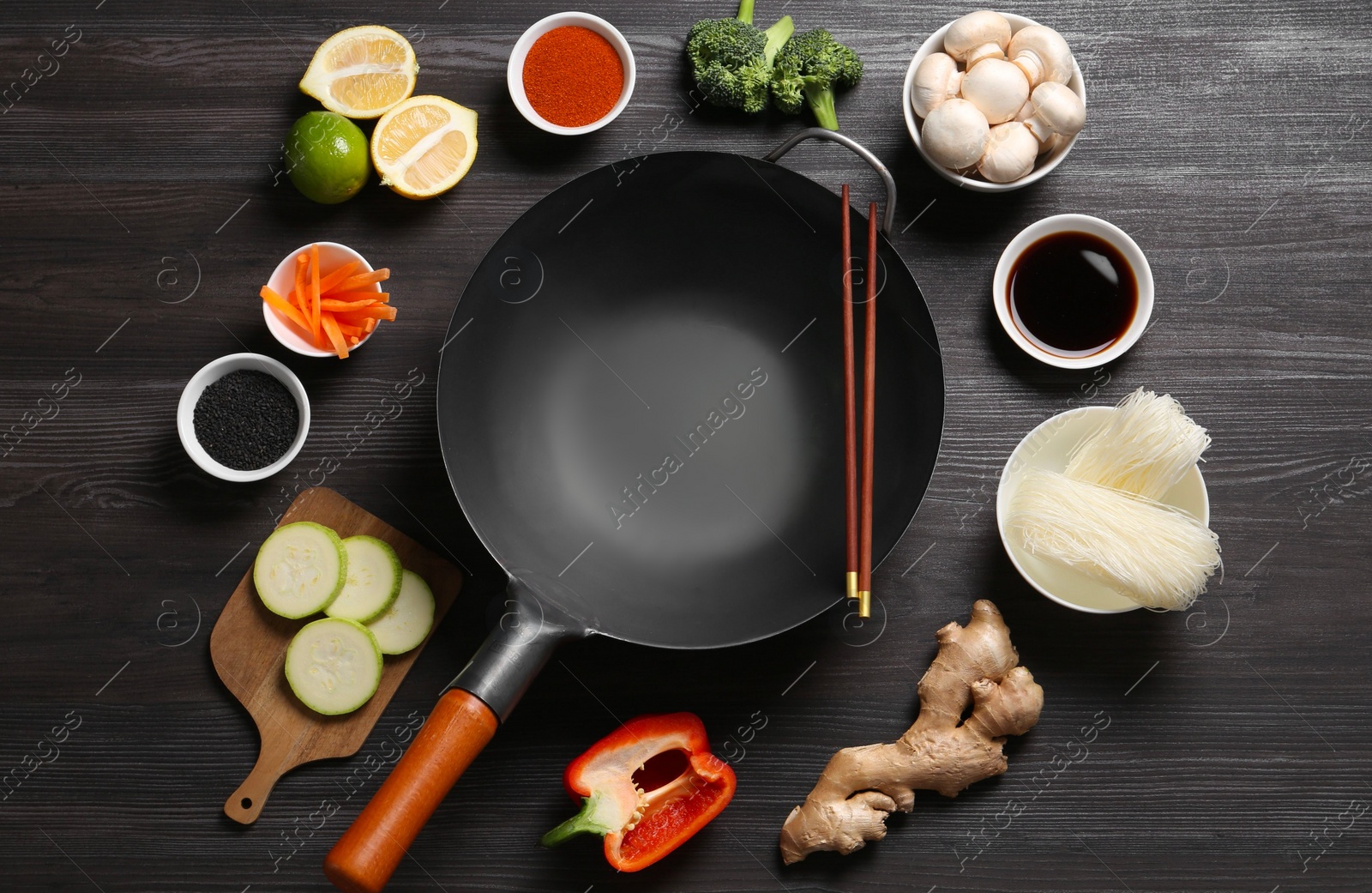 Photo of Empty iron wok and chopsticks surrounded by ingredients on dark grey wooden table, flat lay