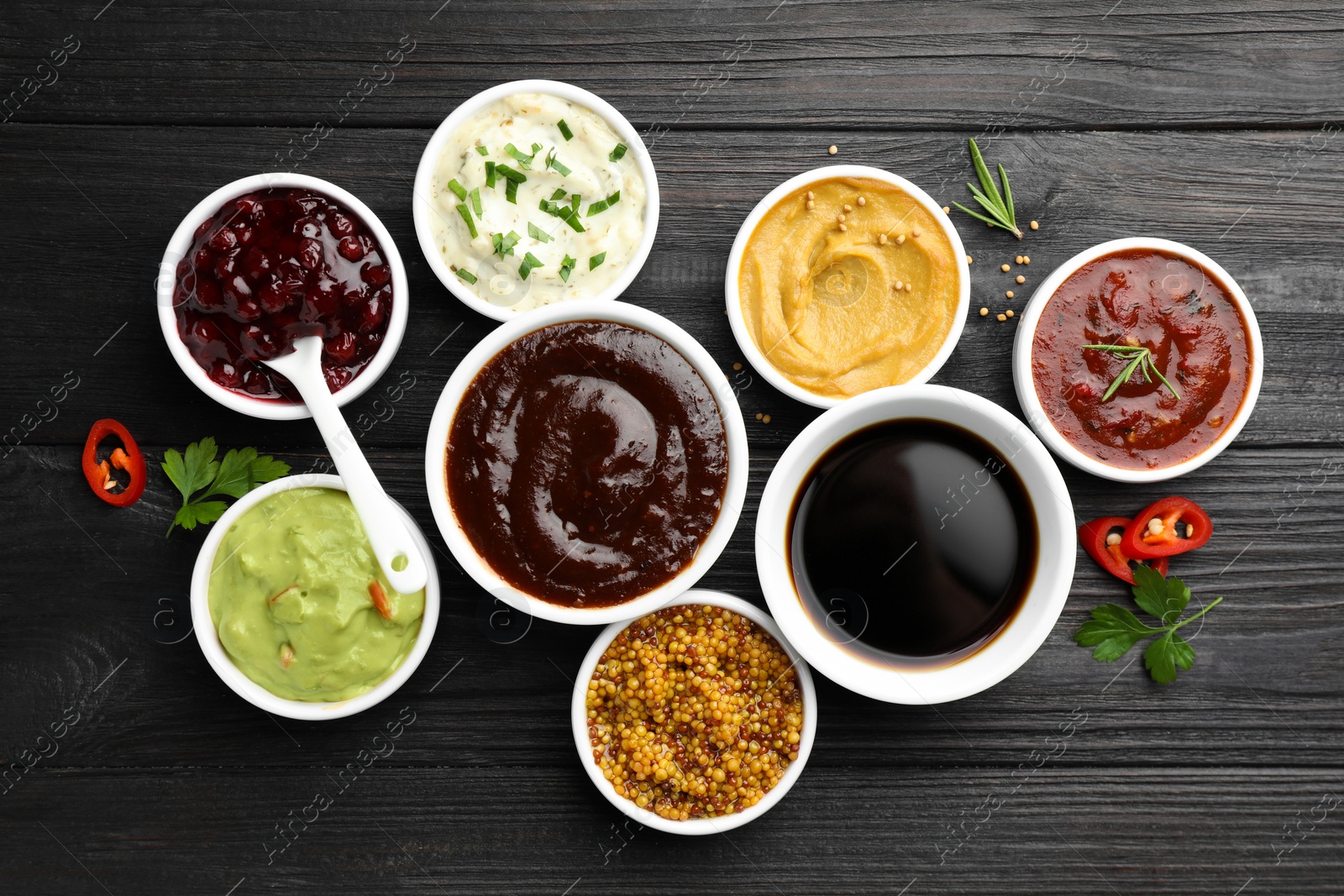 Photo of Different tasty sauces in bowls, parsley, chili pepper and rosemary on black wooden table, flat lay