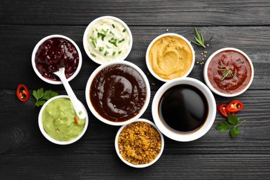 Different tasty sauces in bowls, parsley, chili pepper and rosemary on black wooden table, flat lay