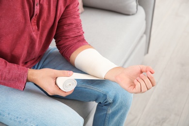 Photo of Young man applying bandage on injured arm at home, closeup. First aid