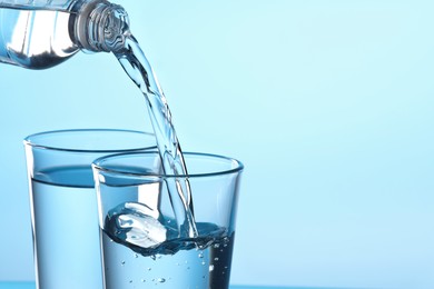 Photo of Pouring water from bottle into glass on light blue background, closeup. Space for text