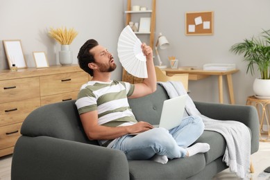 Bearded man with laptop waving white hand fan to cool himself on sofa at home